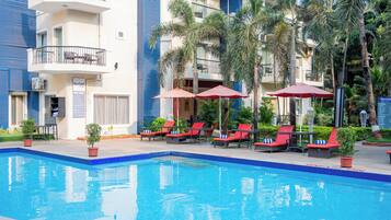 Una piscina con sillas de descanso rojas y sombrillas, rodeada de palmeras y un edificio de varios pisos con balcones.