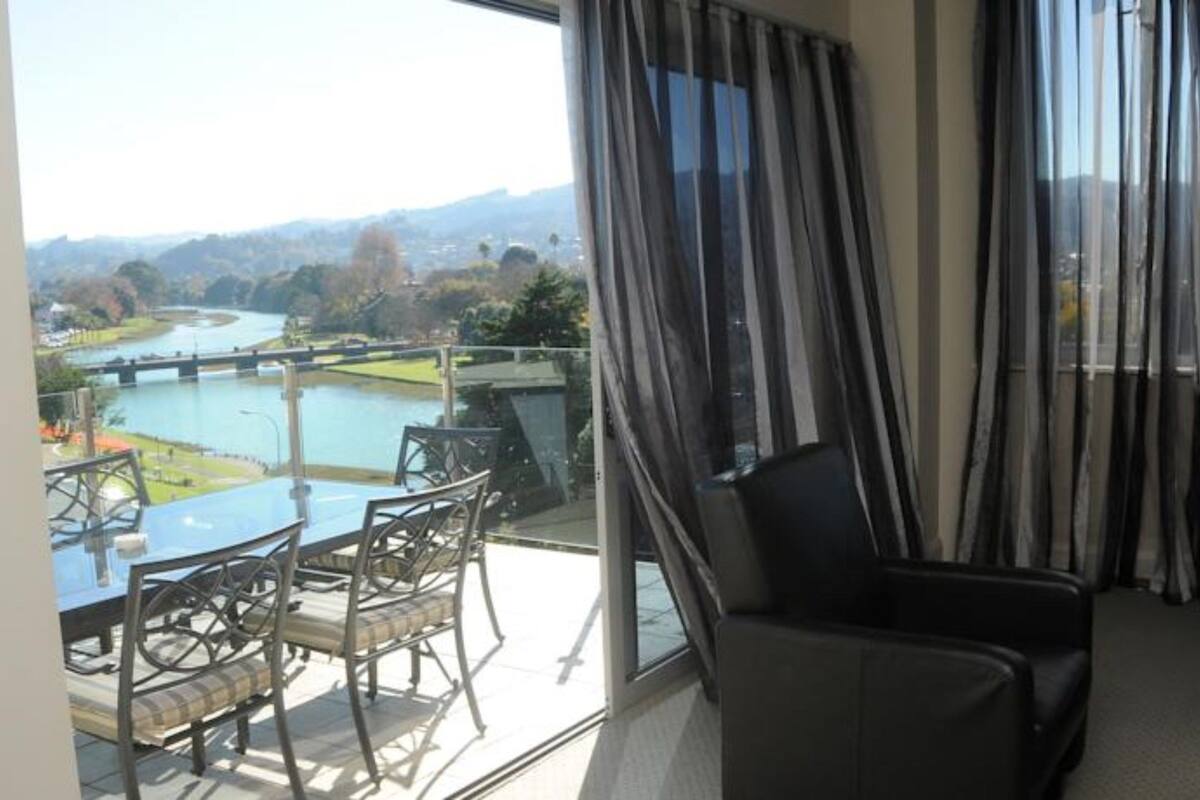 A balcony with a view of a river and bridge, a dining table with chairs, and a black leather chair.