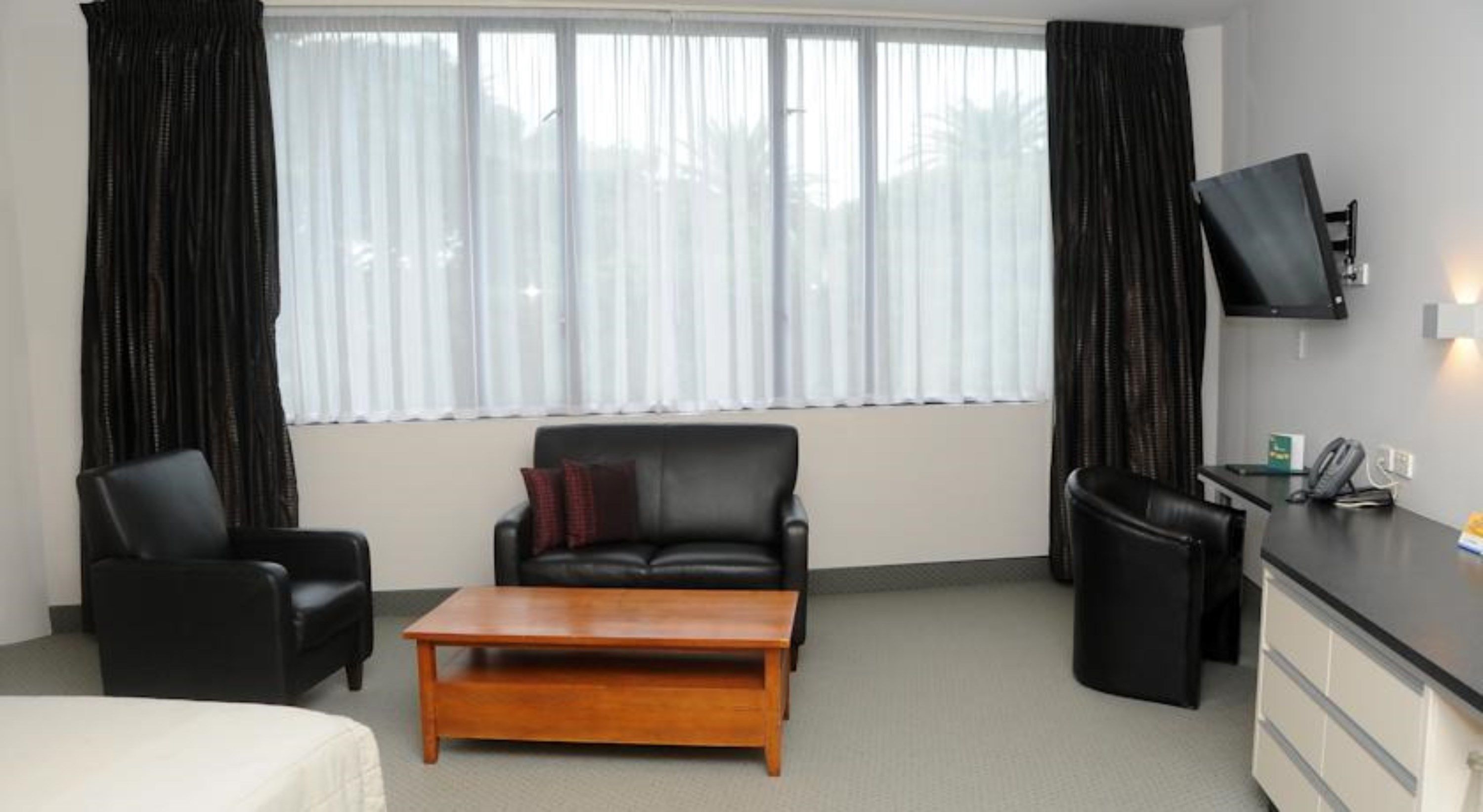 Habitación de hotel con sofá de cuero negro, una mesa de centro de madera, un sillón negro, un escritorio con teléfono y un televisor montado en la pared.