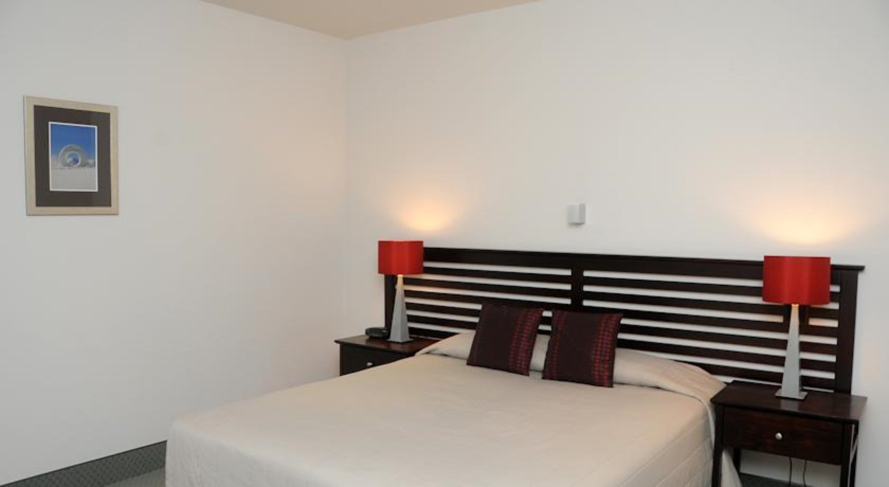 A neatly made bed with a white bedspread, two red lamps on either side, and a framed picture on the wall.