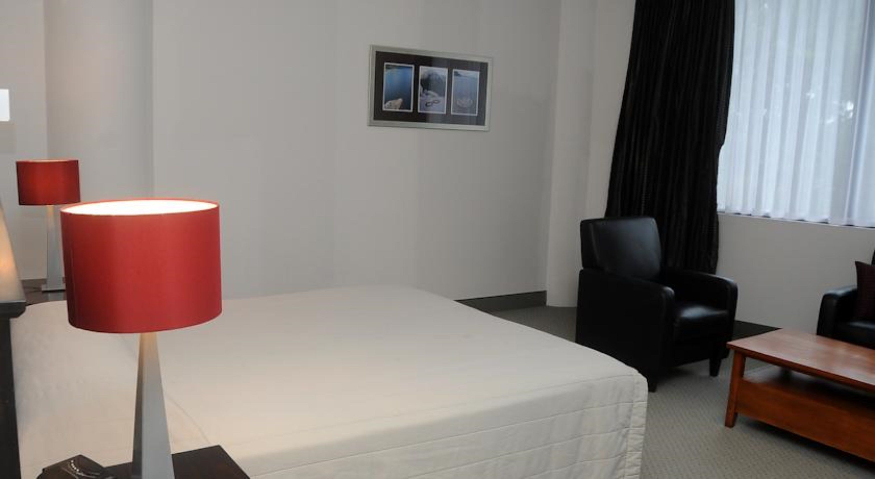 A hotel room with a bed, a desk, a chair, a red lamp, and a picture on the wall.