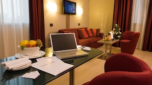 A hotel room with a glass desk, a laptop, a bowl of fruit, a red armchair, a sofa, and a small table with a vase of flowers. - Hotel Cosmopolitan Bologna (Bologna)
