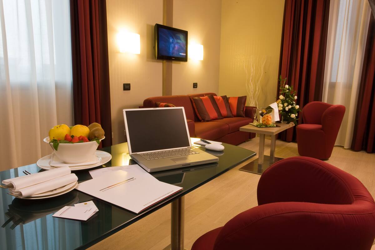 Habitación de hotel con un escritorio de vidrio, una computadora portátil, un cuenco de fruta, un sillón rojo, un sofá y una mesita con un jarrón de flores.