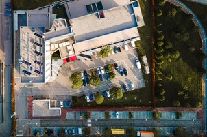 Vista aérea de un estacionamiento con varias filas de coches aparcados, un área peatonal central y un edificio de techo plano.