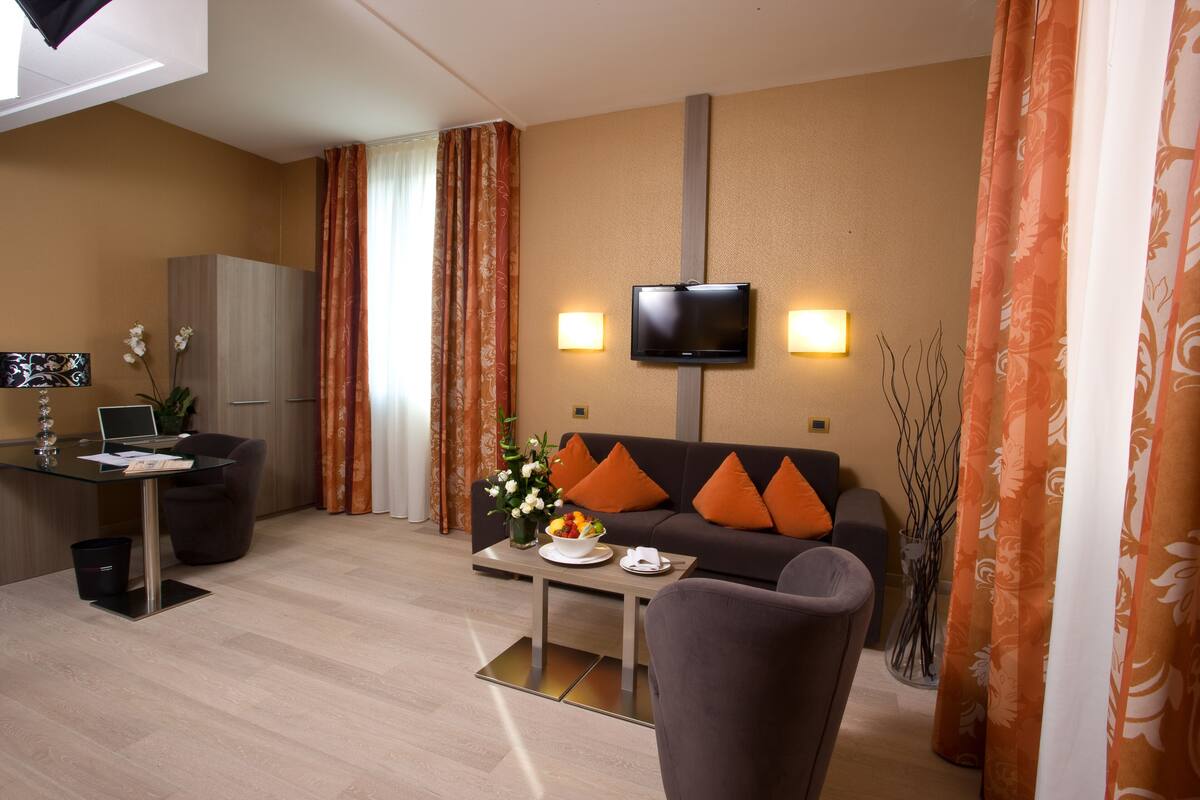 Una habitación de hotel moderna con sofá, una mesa de comedor, un televisor montado en la pared y un escritorio con una computadora portátil.
