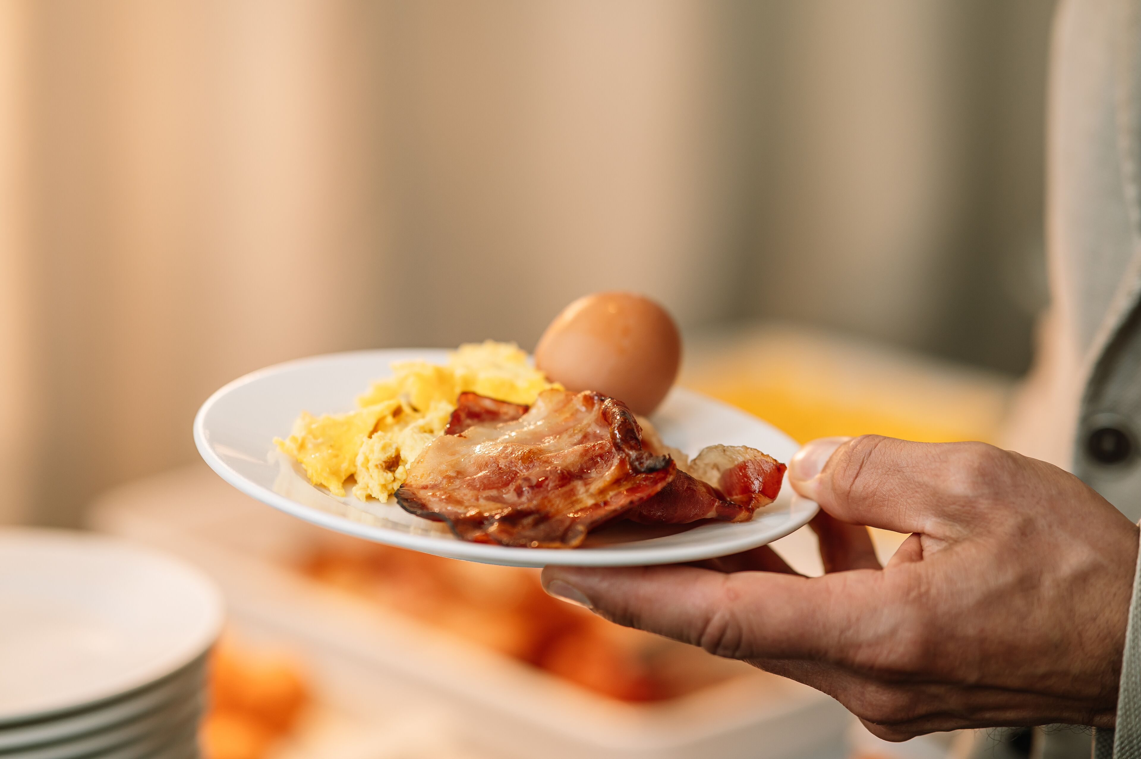 A person holding a plate with scrambled eggs, bacon, and a boiled egg.