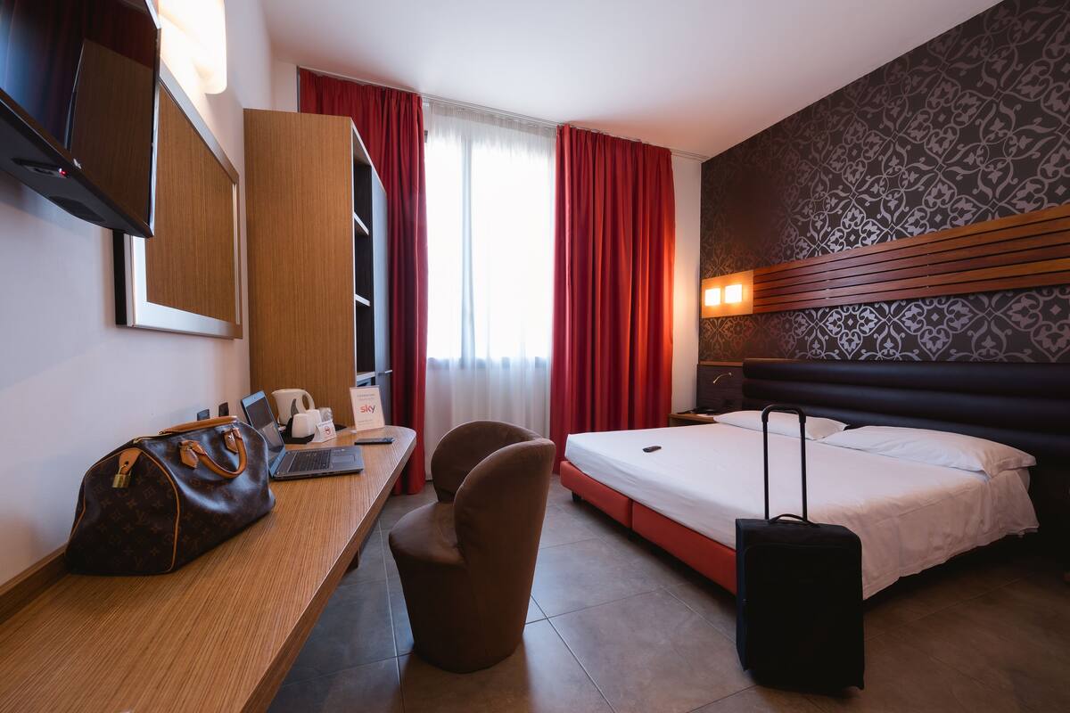 Habitación de hotel con cama, escritorio con computadora portátil, silla, maleta, armario y un espejo grande.
