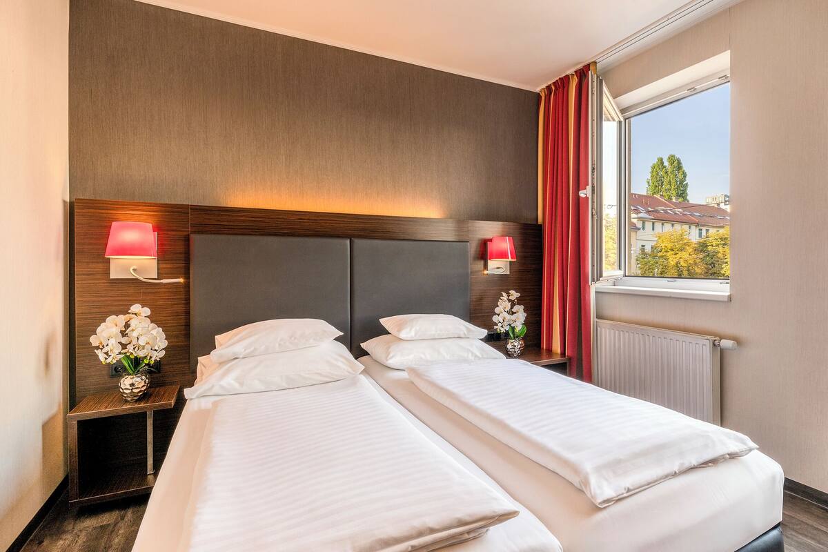 Une chambre d’hôtel avec deux lits, une tête de lit en bois, des lampes de chevet, une fenêtre donnant sur des immeubles et un fauteuil rouge.
