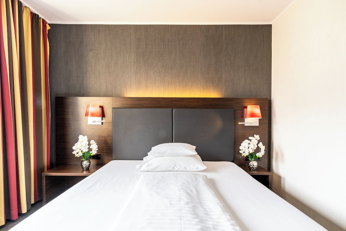 Une chambre d’hôtel avec un grand lit, deux tables de chevet avec des lampes, une tête de lit dotée d’un éclairage intégré et un rideau à rayures verticales.