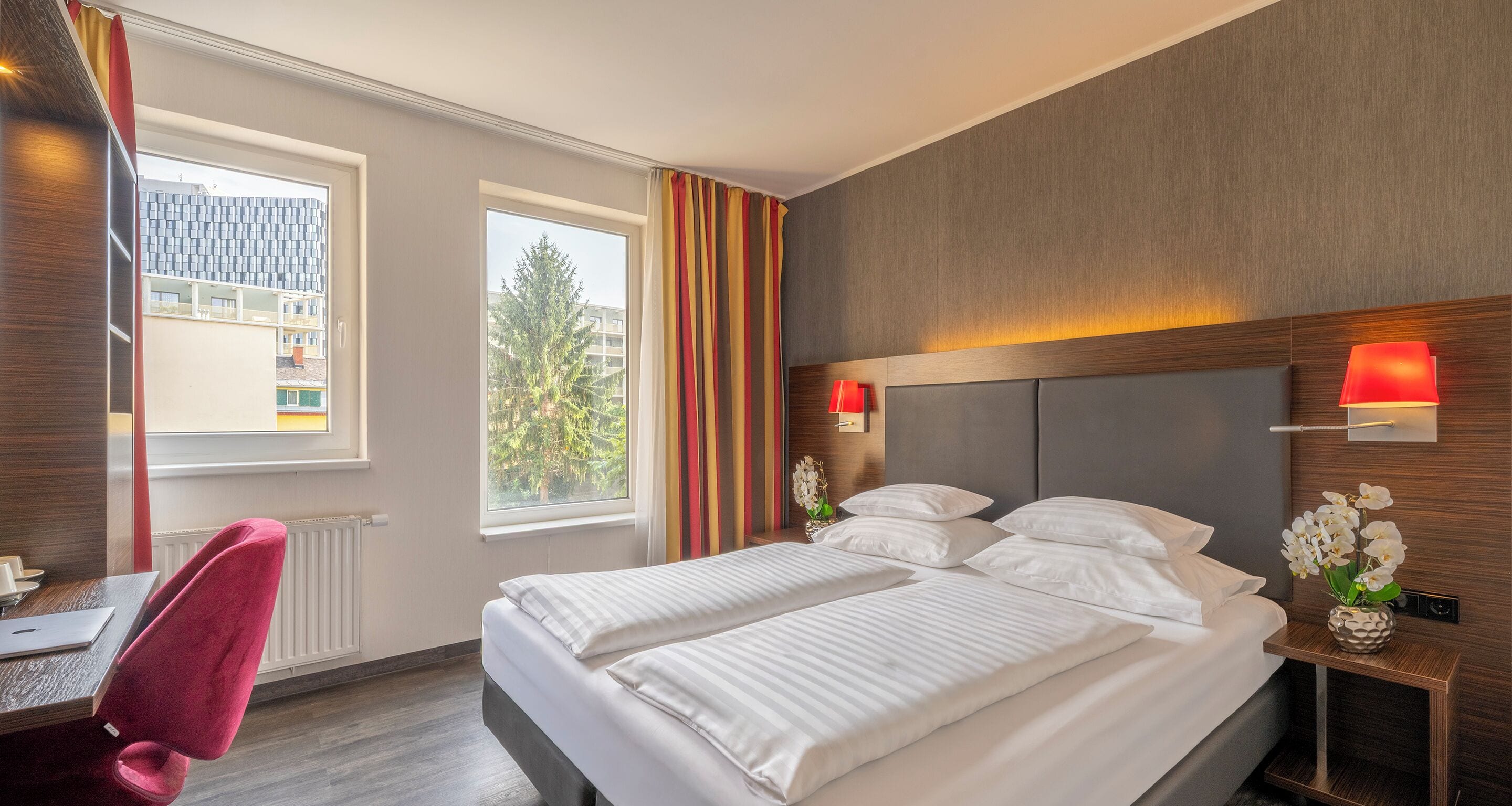 A hotel room with a double bed, a desk with a chair, a window with curtains, and a view of buildings outside.