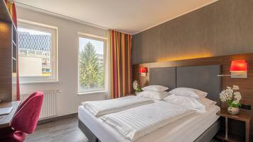 A hotel room with a double bed, a desk with a chair, a window with curtains, and a view of buildings outside.
