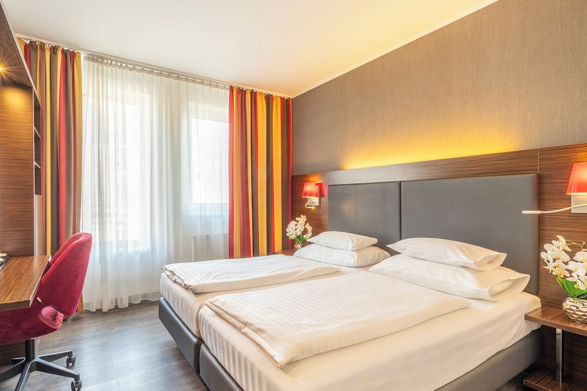Une chambre d’hôtel avec deux lits, une chaise rouge, une table de chevet avec une lampe et une fenêtre avec des rideaux.
