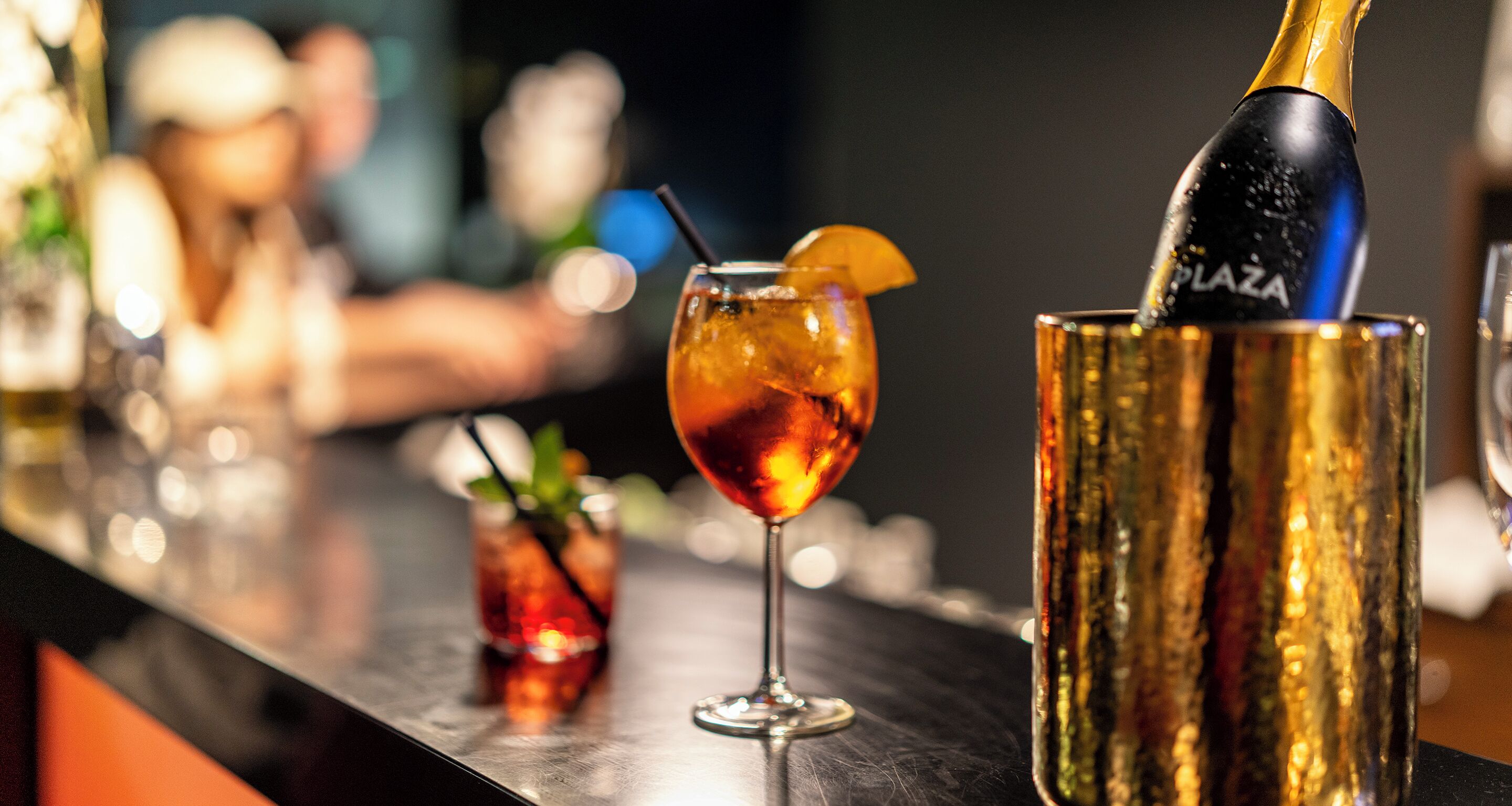 A bar setting with a champagne bottle being poured into a glass, a cocktail with an orange garnish, and a small glass with a green garnish.