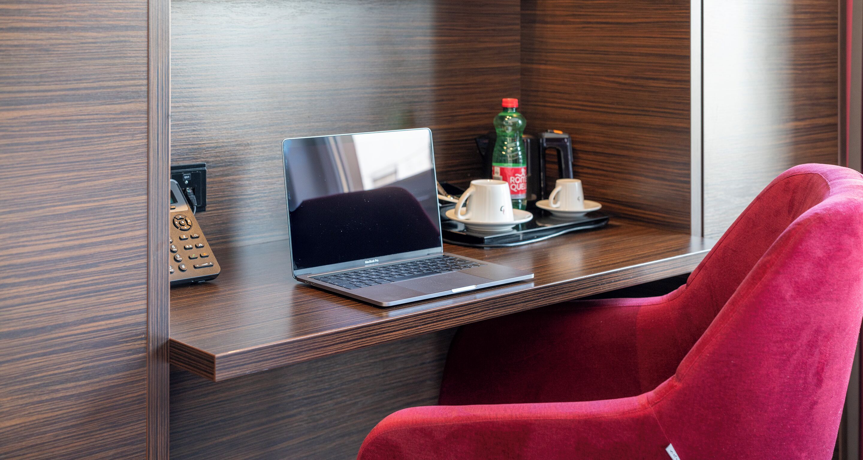 Una habitación de hotel moderna con un escritorio de madera, una computadora portátil, una silla roja, una cafetera y una botella de agua.