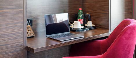 Una habitación de hotel moderna con un escritorio de madera, una computadora portátil, una silla roja, una cafetera y una botella de agua.