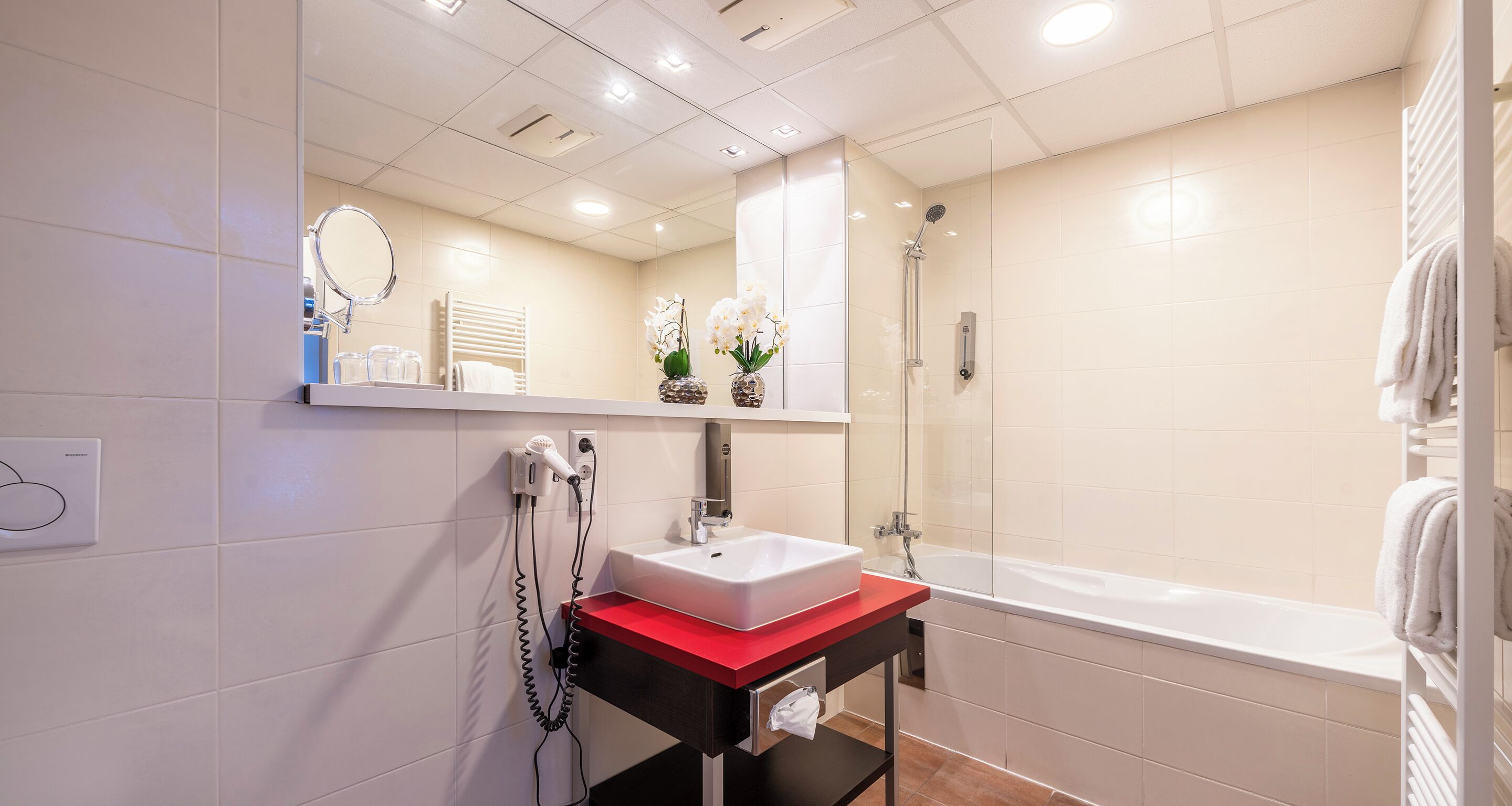 Un baño moderno con un lavabo blanco, una encimera roja, una ducha con mampara de cristal y un espejo sobre el lavabo.