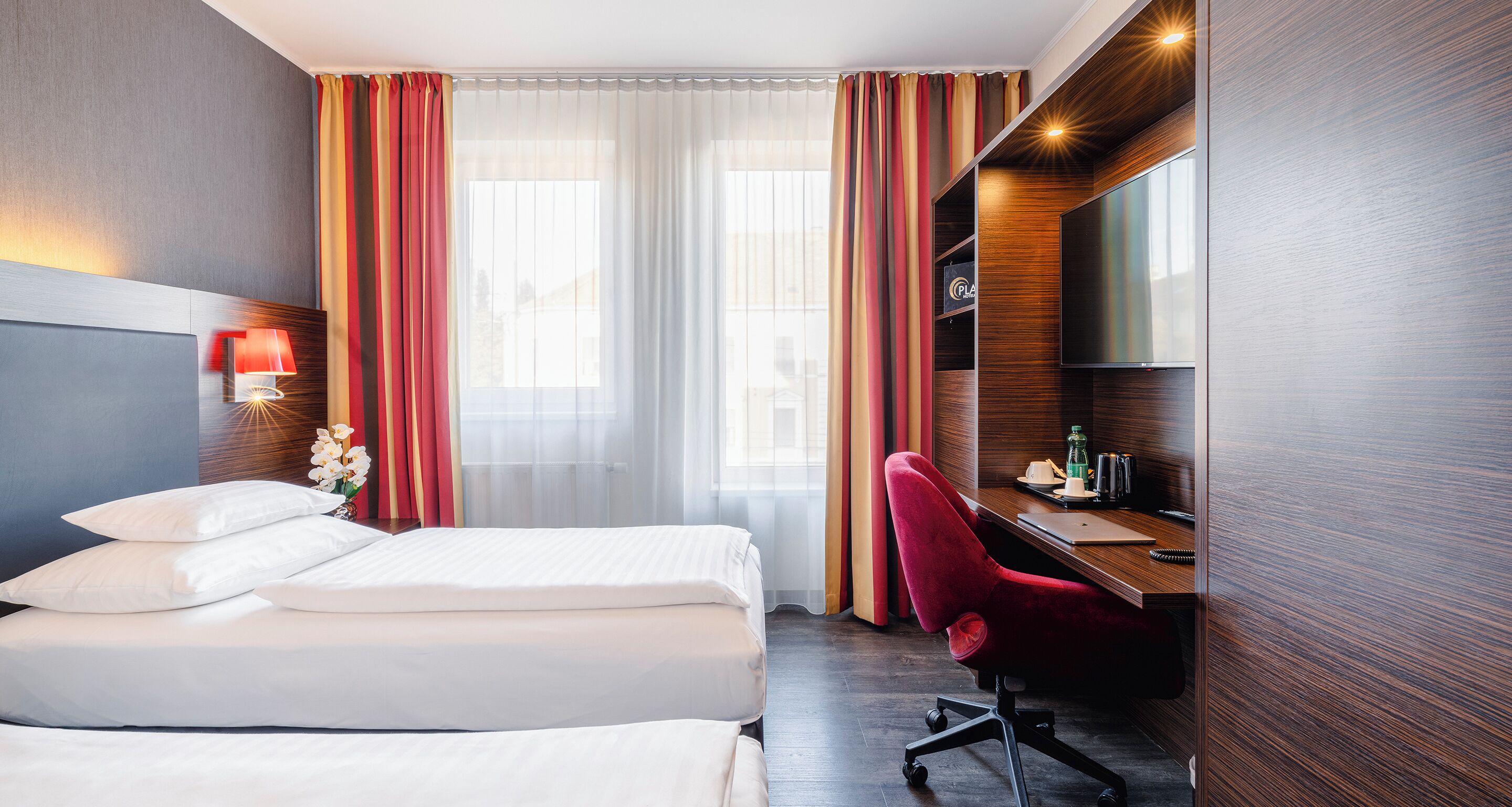 A hotel room with two beds, a desk with a chair, a TV, and a window with curtains.