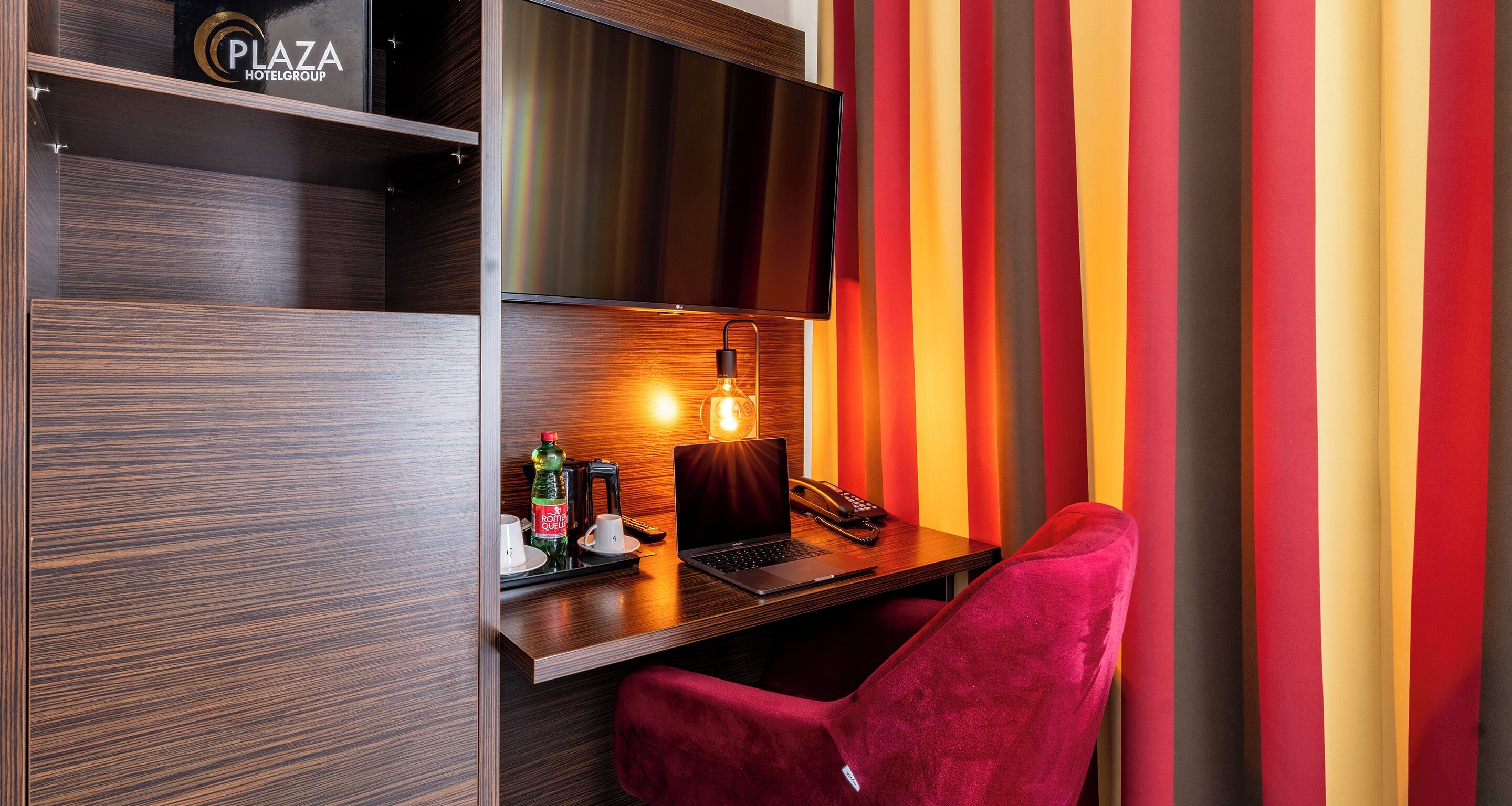 A hotel room with a wooden desk, a laptop, a red chair, a minibar, and a TV on a wooden shelf.