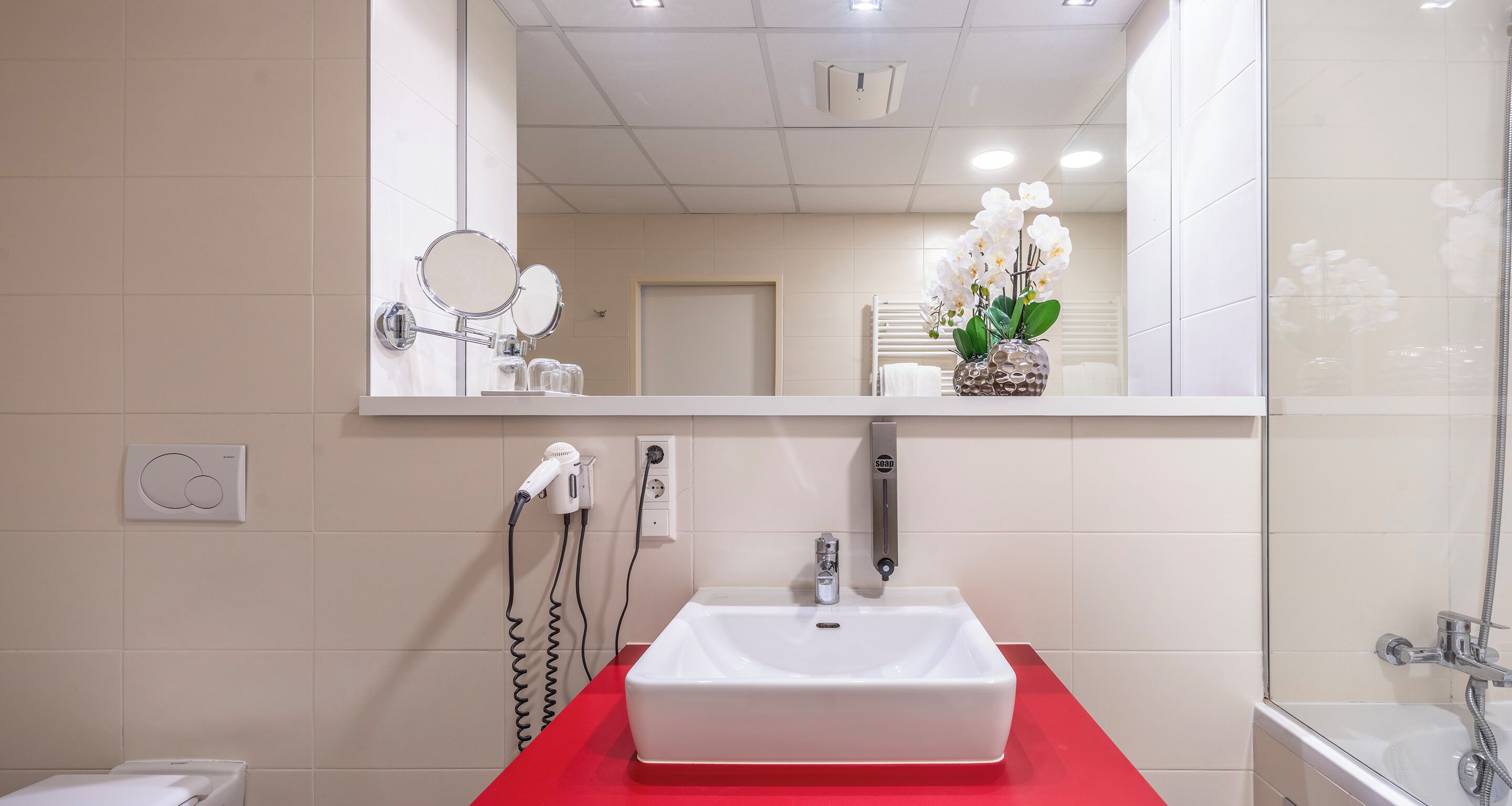 A bathroom with a white sink, red countertop, and a mirror above it. There is a hairdryer and a soap dispenser on the wall.
