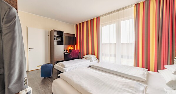 Ein Hotelzimmer mit zwei Betten, einem Schreibtisch mit Computer, einer Minibar und einem Fenster mit Vorhängen.