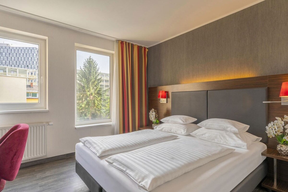 Una habitación de hotel con cama, dos lámparas de noche, un escritorio con silla y una ventana con cortinas.