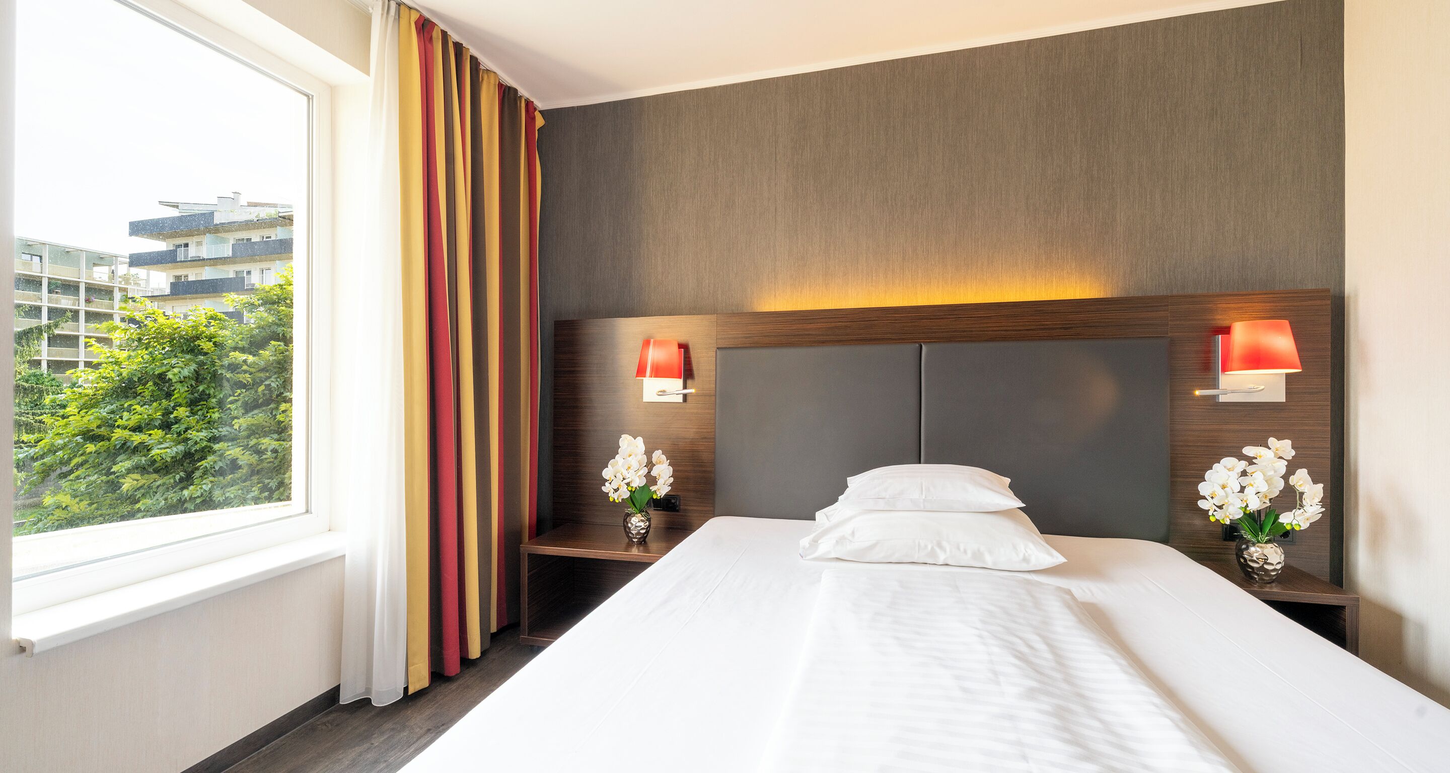 A hotel room with a large bed, two bedside tables with lamps, a window with curtains, and a view of buildings and trees outside.