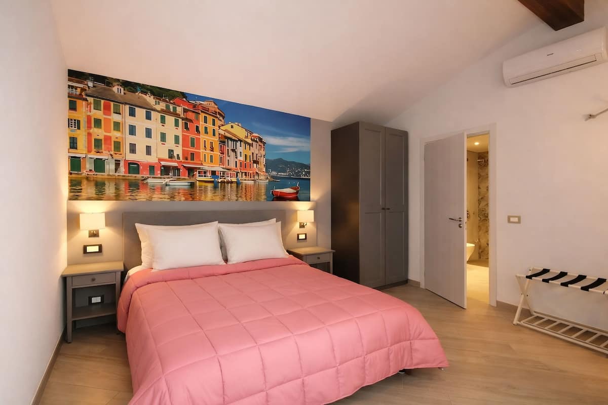 Un dormitorio con cama, mesitas de noche, armario, un cuadro de un pueblo costero y la puerta del baño.
