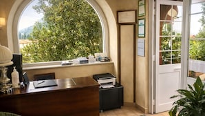 A reception desk with a view of trees and mountains through a large window. There is a door to the right, and a potted plant is visible in the foreground. - Hotel Del Borgo (Sestri Levante)