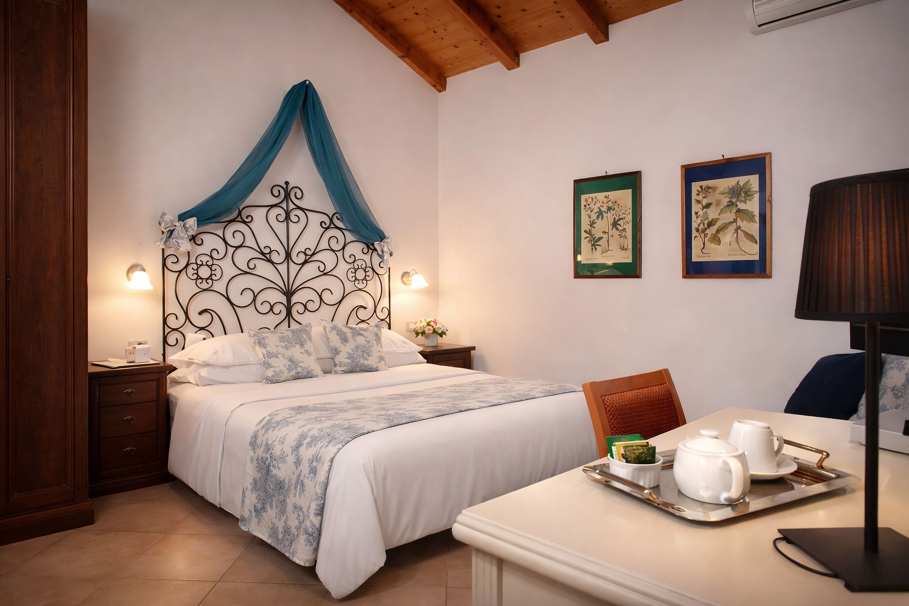 Un dormitorio ordenado con una cama, mesitas de noche, un escritorio con juego de té e impresiones botánicas enmarcadas en la pared.