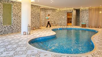 Uma grande piscina interior com paredes e chão em mosaico, rodeada por um tapete estampado.
