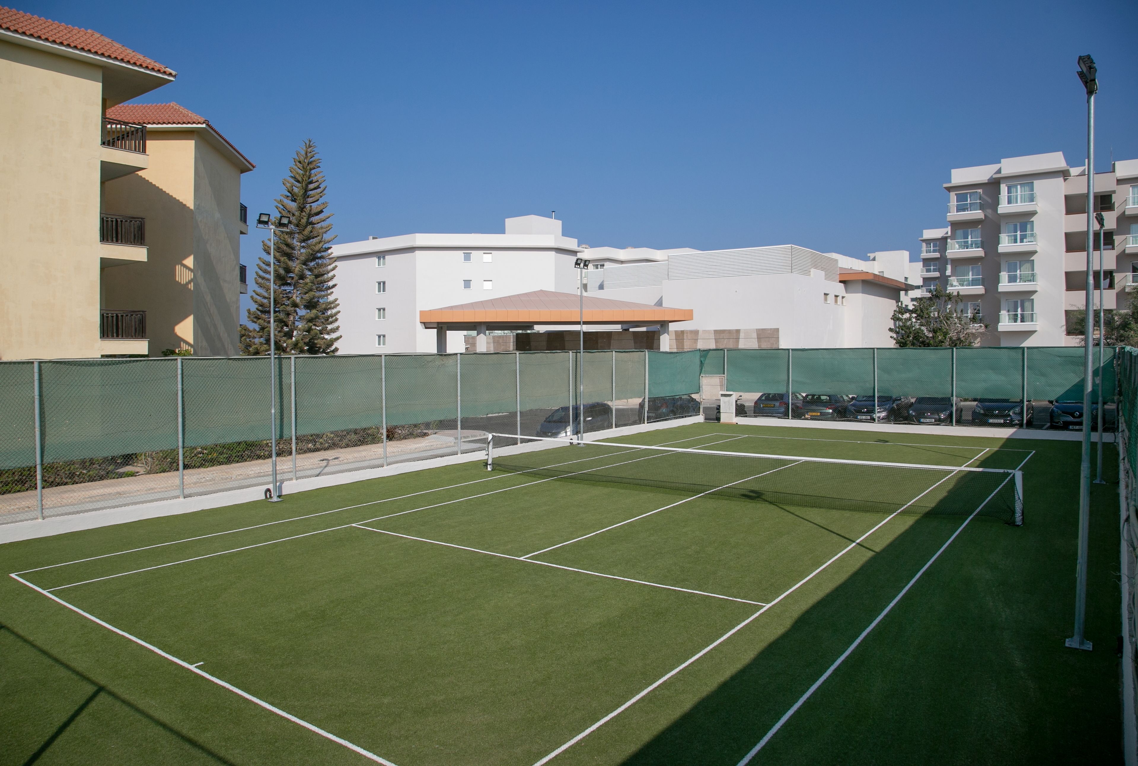 An outdoor tennis court with a net, surrounded by a green fence, and adjacent to residential buildings.
