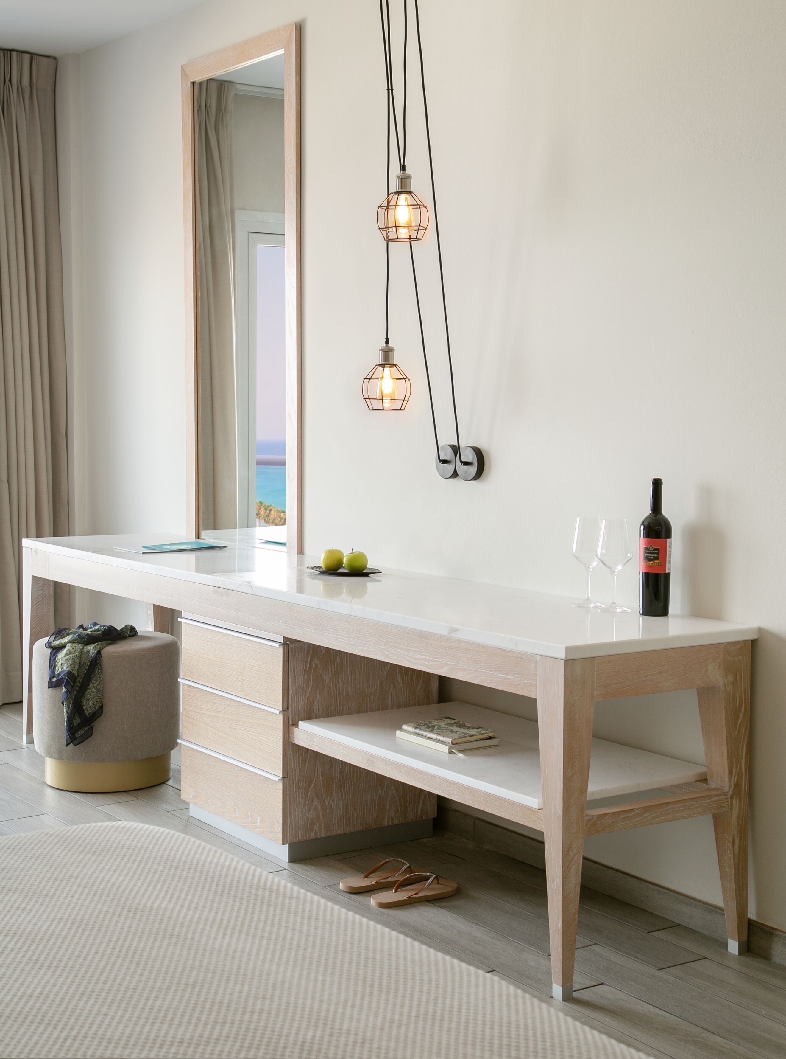 A modern bedroom with a large mirror, a wooden desk with drawers, a hanging light fixture, and a bottle of wine on the desk.