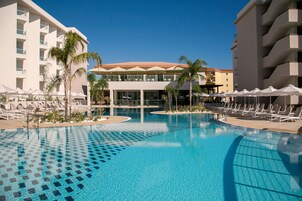 A hotel pool area with multiple lounge chairs and umbrellas, surrounded by palm trees and a building with balconies.