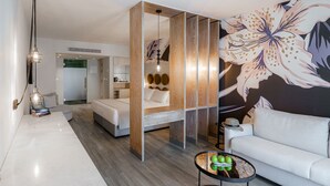 Una habitación de hotel moderna con una cama grande, un sofá, una mesita redonda con manzanas y una pared decorativa con motivos florales.