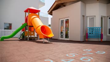 A playground with a slide and a climbing structure, a blue and orange playhouse, and a building with a white exterior and a door with glass panels.