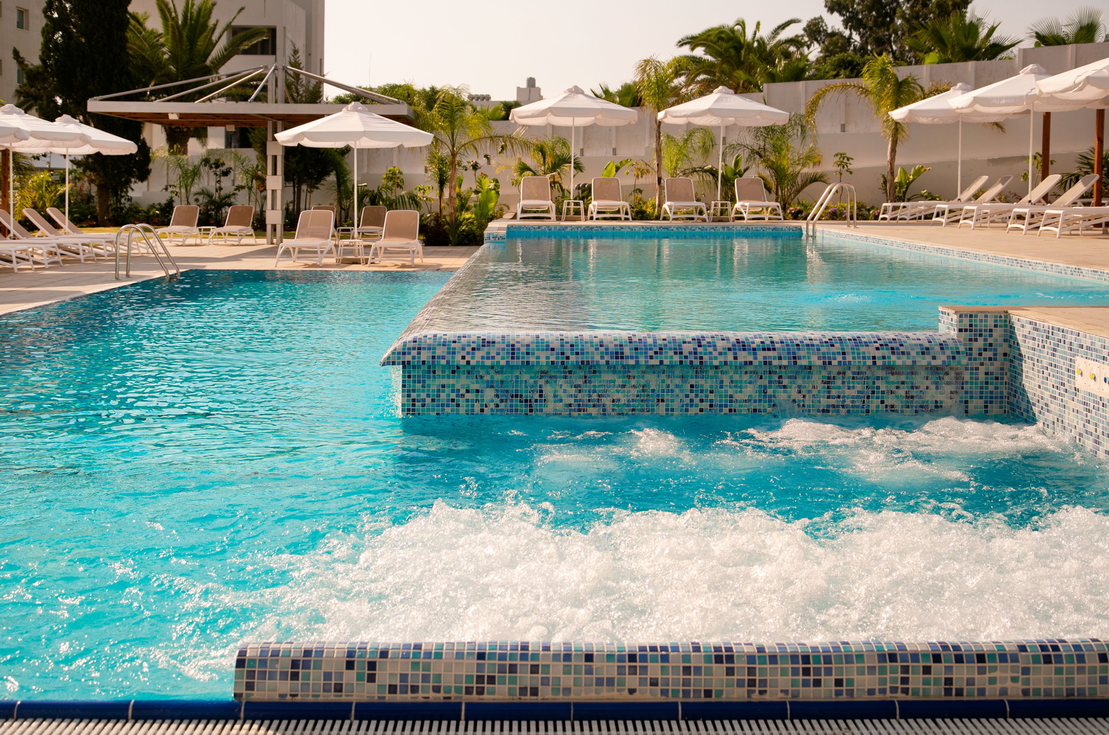 Ein Pool mit einer Fliesenleiste, umgeben von Liegestühlen und Sonnenschirmen.