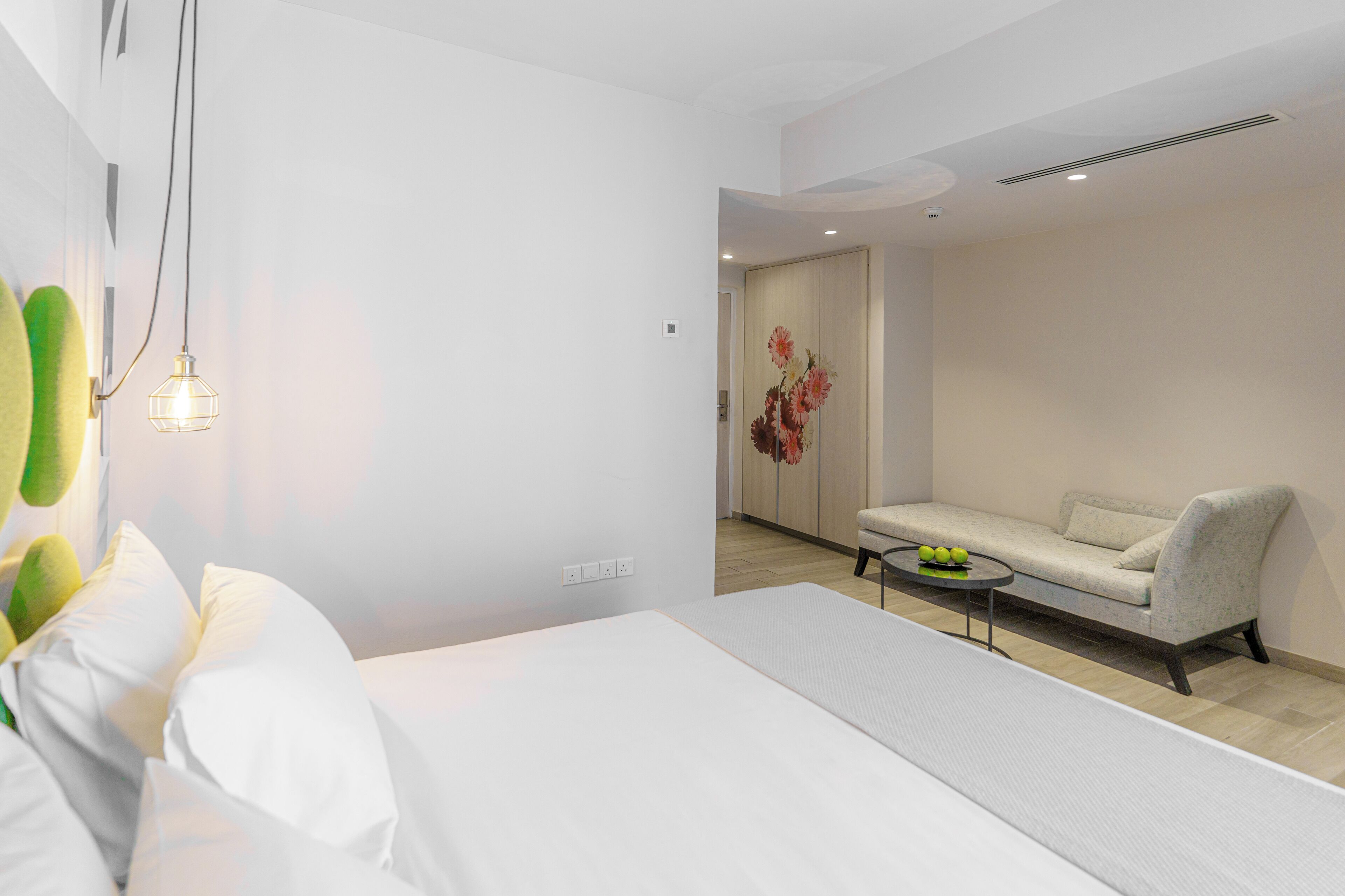 A modern hotel room with a large bed, a sofa, a small table with fruit, and a decorative flower arrangement.