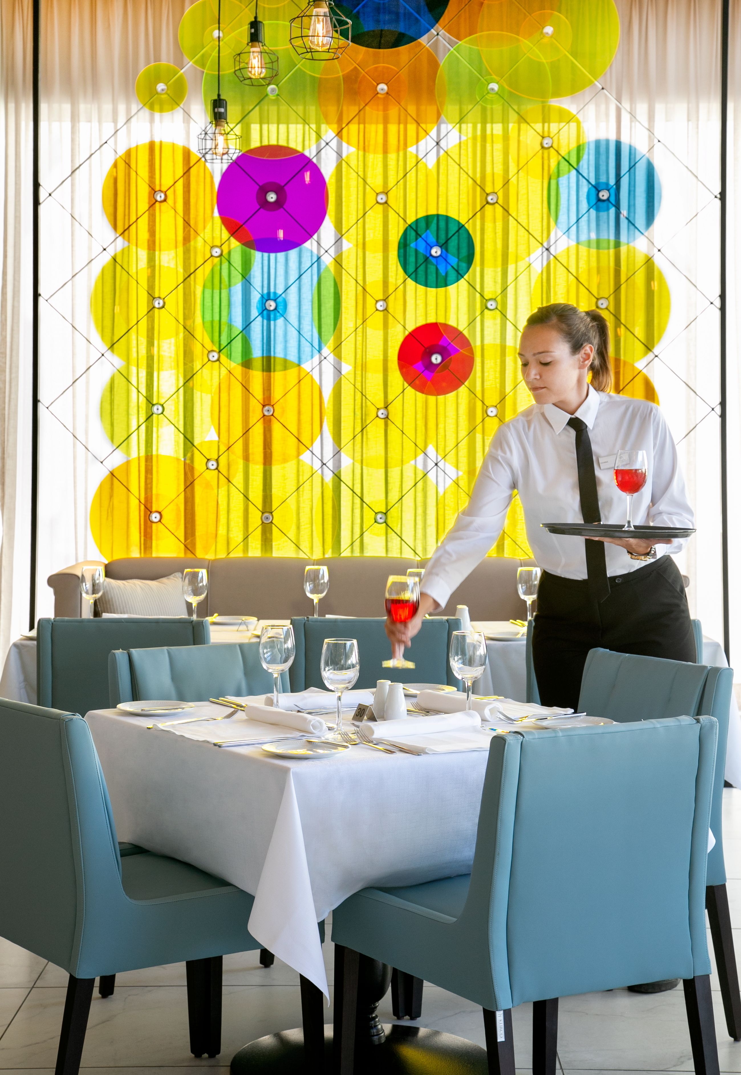 A dining area with a table set for two, a waiter serving wine, and a colorful abstract wall in the background.