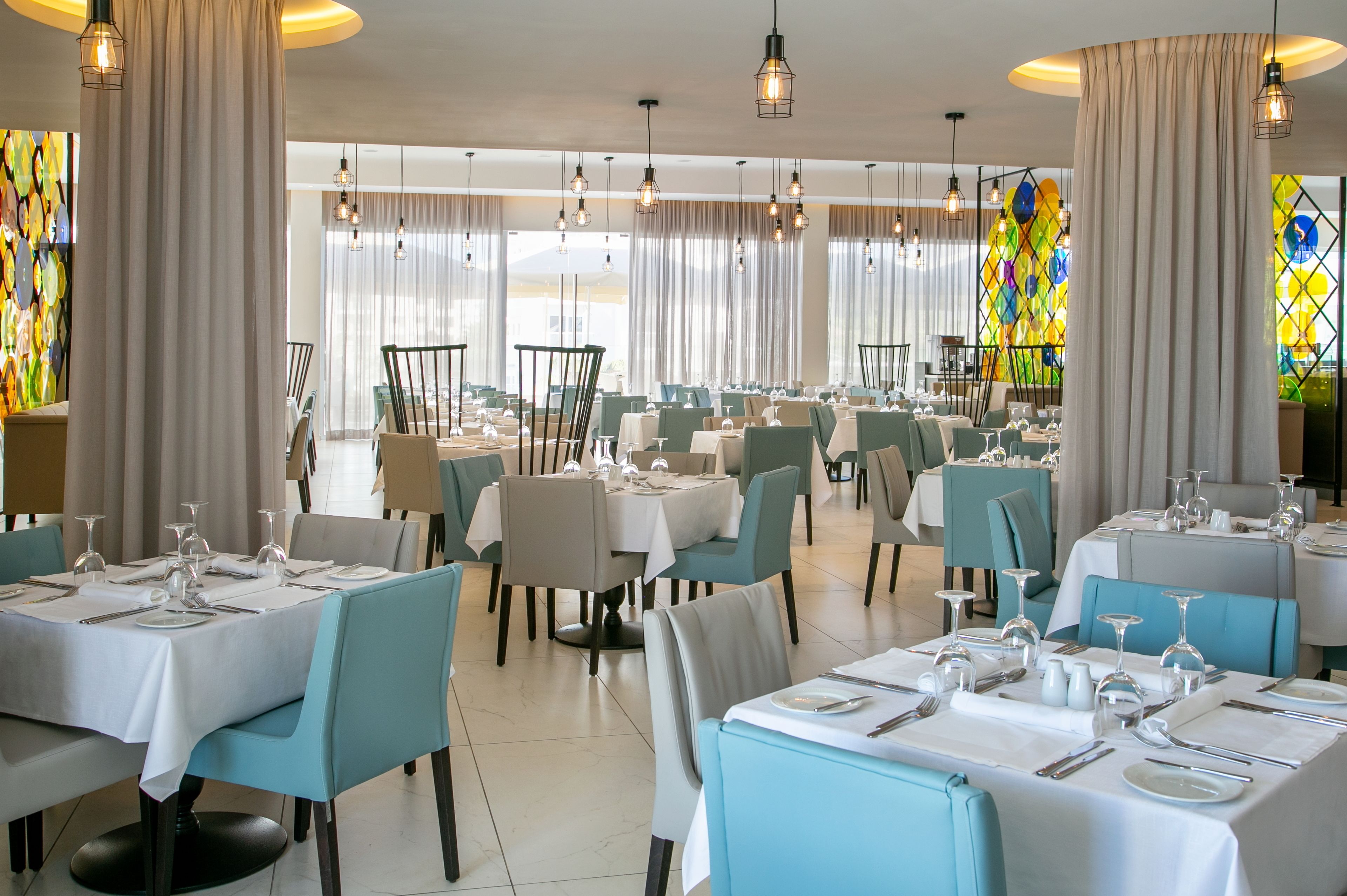 Un restaurante con varias mesas redondas dispuestas para comer, con sillas azul claro y manteles blancos.