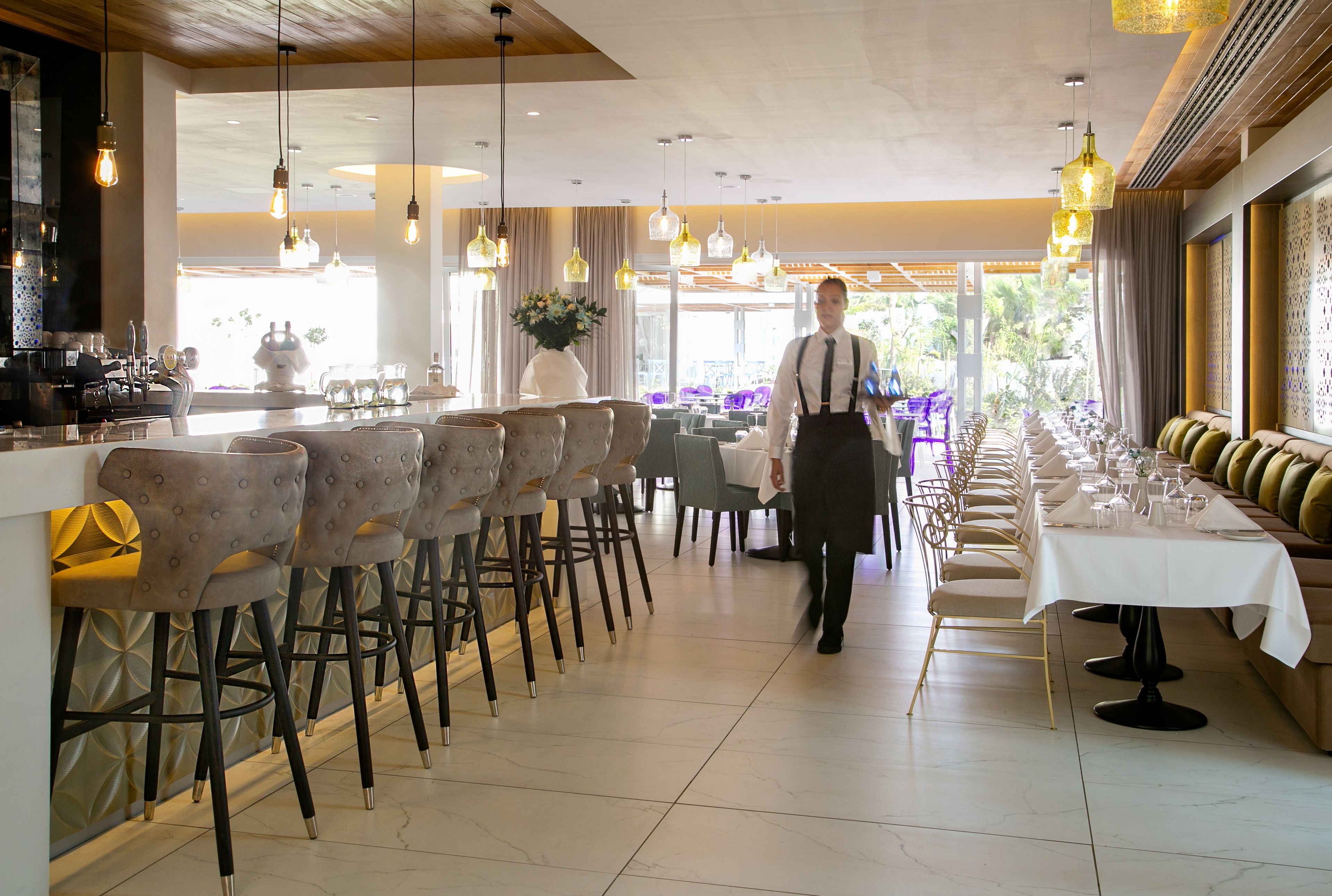 A restaurant with a long dining table, multiple chairs, and hanging pendant lights.