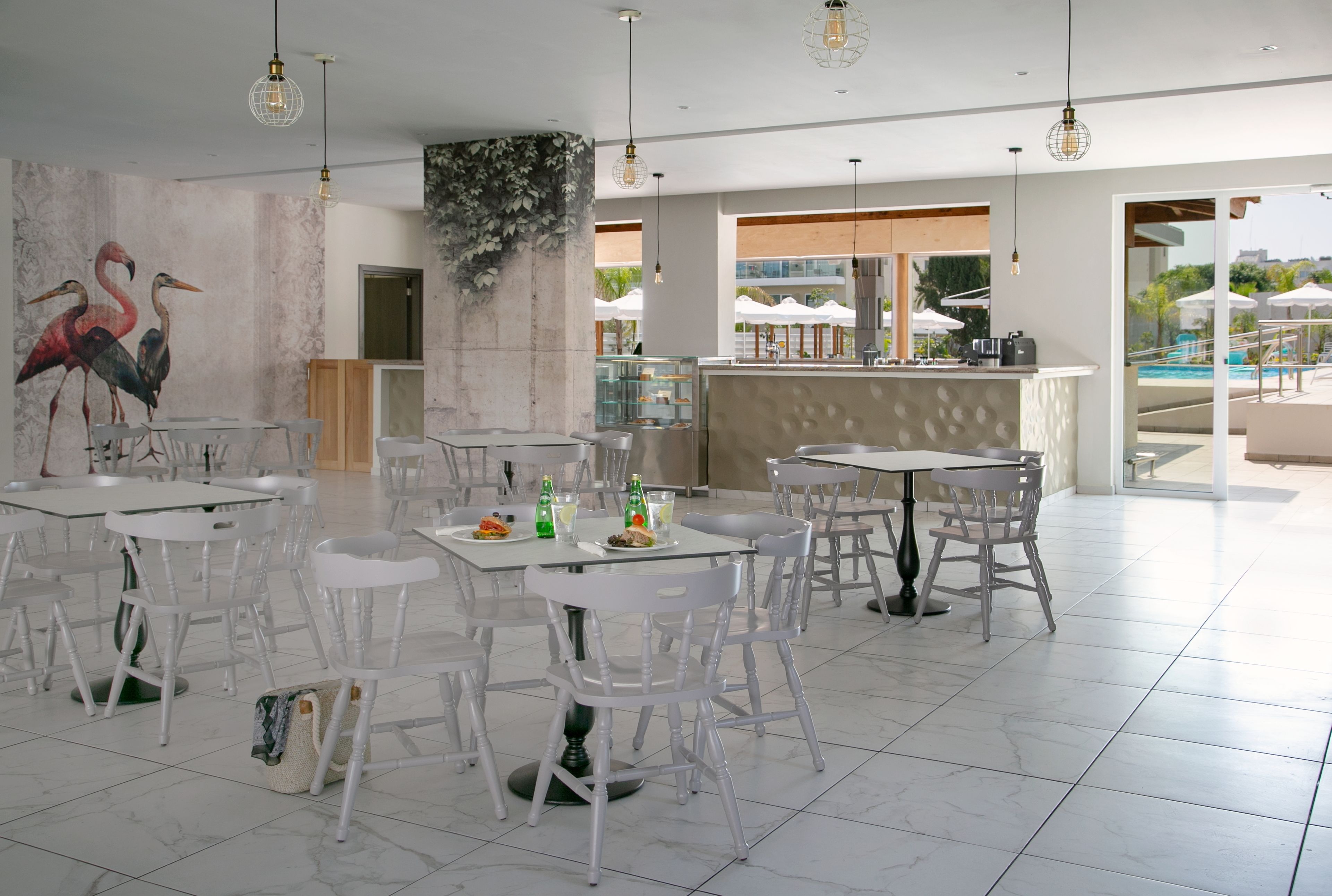 A spacious dining area with white tables and chairs, a mural of flamingos, and a bar area with a view of the pool.