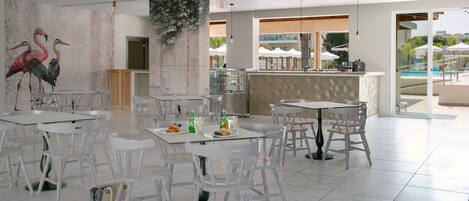 A spacious dining area with white tables and chairs, a mural of flamingos, and a bar area with a view of the pool.