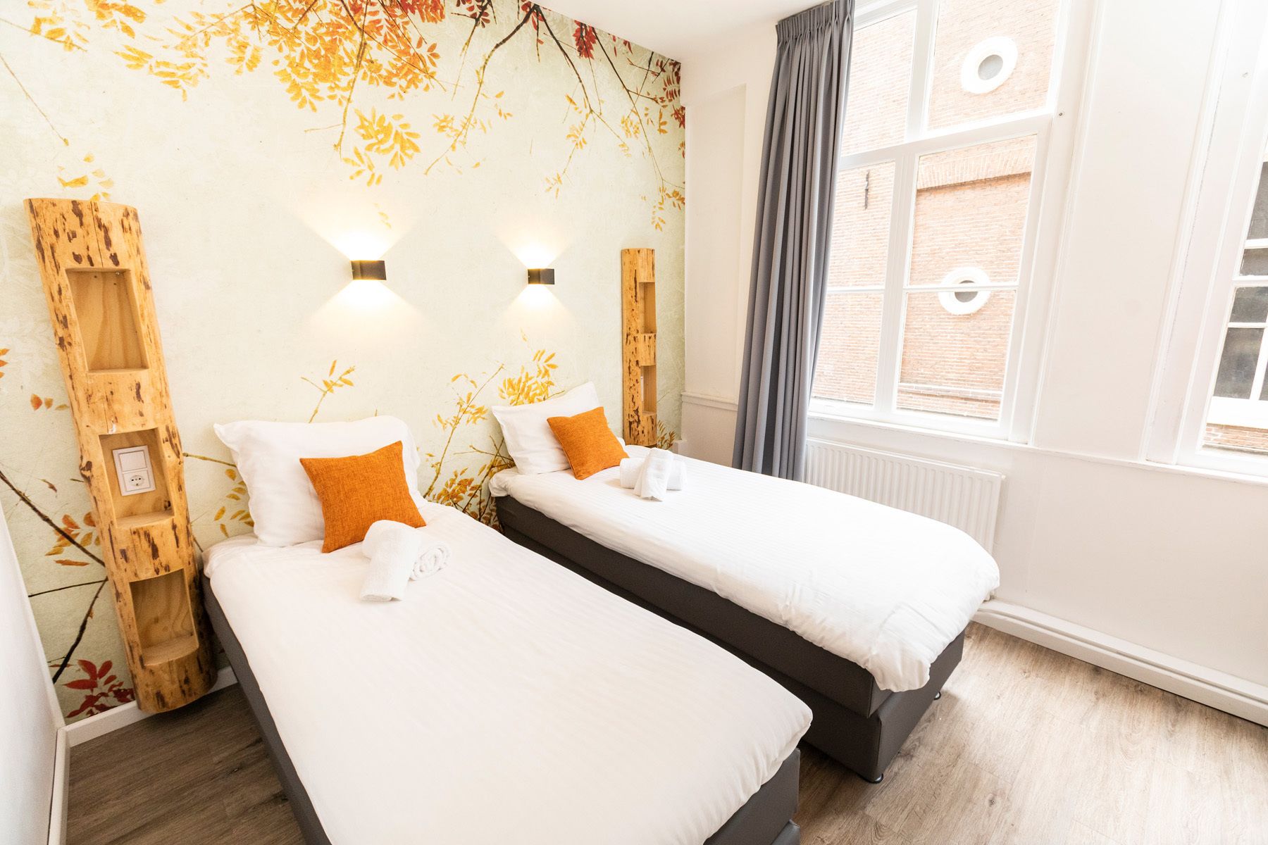 A hotel room with two beds, a wooden ladder, a wall with autumn leaves, and a window with curtains.