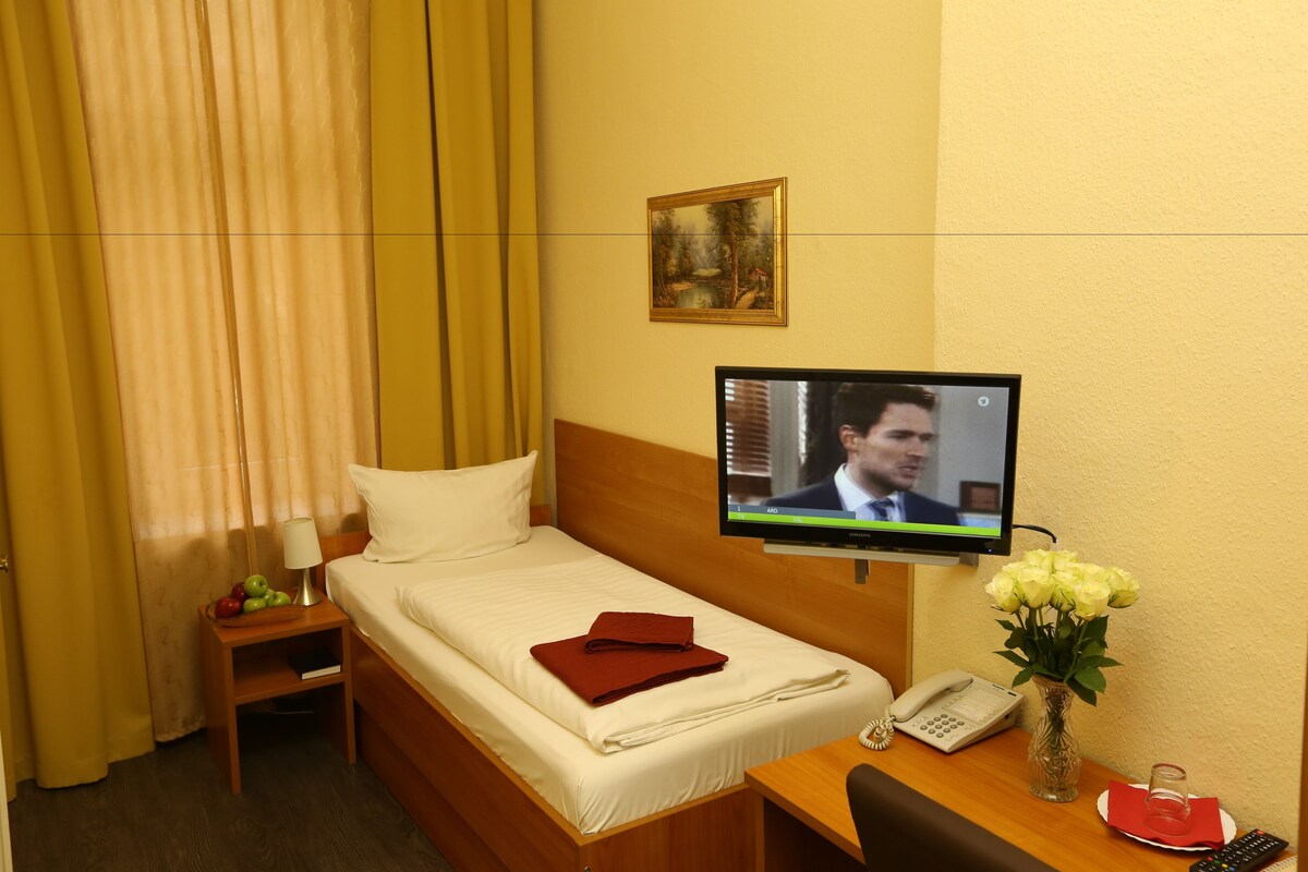 A hotel room with a bed, a desk with a phone, a vase with flowers, and a television on the wall.