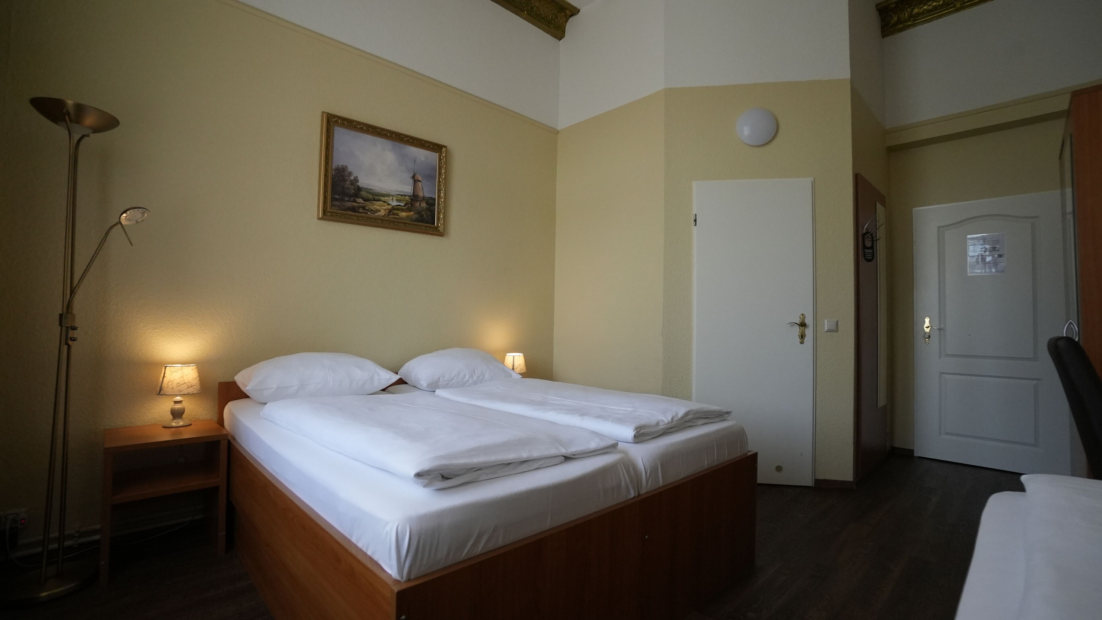 A room with a bed, bedside lamps, a painting, a door, and a lamp on the floor.