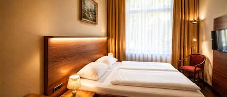A hotel room with a wooden headboard, a bed with white linens, a bedside table with a lamp, a red chair, and a television on the wall.