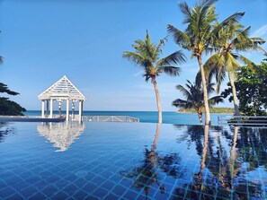 Une piscine à débordement avec un belvédère blanc et des palmiers, offrant une vue imprenable sur une mer d’un bleu profond.