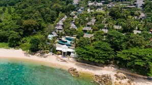 An aerial view of a beach resort with a clear turquoise sea, a sandy beach, and a cluster of thatched-roof cottages surrounded by dense greenery. - Bhundhari Resort & Villas Samui (Koh Samui)