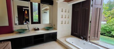 Un baño moderno con un espejo grande, dos lavamanos, una bañera y vistas a zonas verdes al exterior.