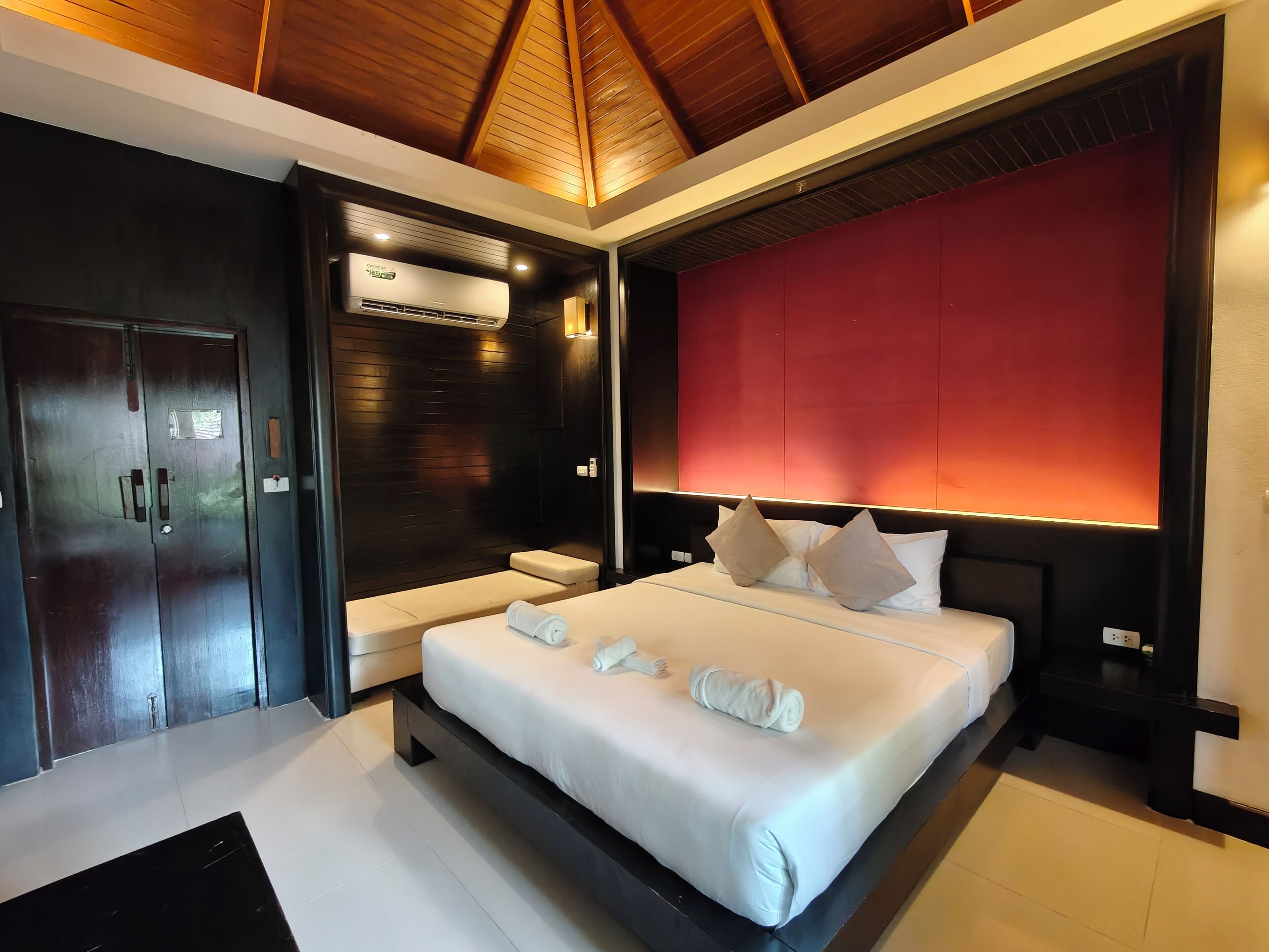 A modern hotel room with a large bed, a built-in bench, and a red accent wall.
